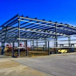 Pre-engineered metal building under construction with exposed steel framing, showing clear-span structure and open worksite on a clear blue day.