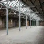 Interior of an empty industrial warehouse with exposed steel beams, brick walls, tall windows, and a sloped skylight roof allowing natural light to enter the large open space.